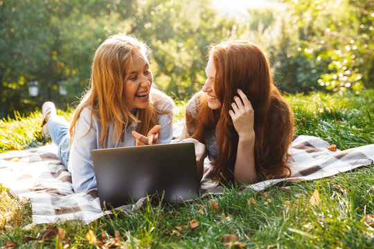 Image of two young women wearing casual clothes lying on grass in park, and using silver laptop