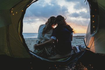 Young couple in love hug each other on the deserted beach on a summer evening at sunset during camping. Point of view inside the tent