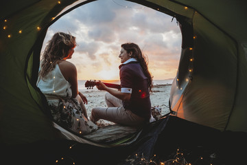 Young loving couple while camping on the seashore a summer evening at sunset. A woman plays the...