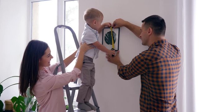 home repair, cute child with measuring tape helps to parents make repairs and hang picture in new apartment
