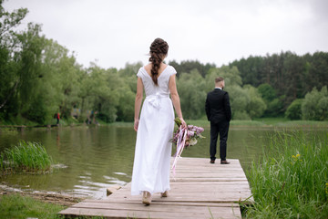 Rear view of young bride woman in beautiful white wedding dress walking to the groom on the pier near the lake. Wedding day.
