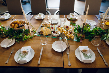 Top view of stylish wedding table setting with empty wine glasses, plates, napkins, green plants and cheese platter on wooden table for rustic wedding. Indoors