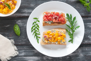 Bruschetta with cheese red and yellow tomatoes on a white plate. The composition is decorated with leaves of basil, arugula, pepper and vegetables. Top view on an old wooden background.