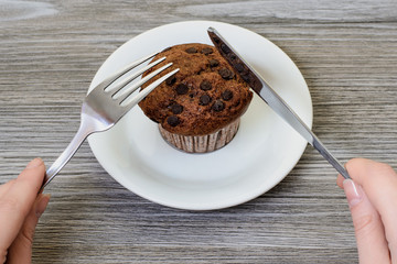 It's brunch time! Woman's hands trying to cut tasty fresh appetising chocolate muffin on white plate. Close up photo.