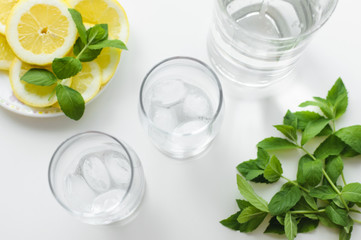 Top view on a beautiful cold drink arrangement, what is two glasses with ice cubes, bottle of water, plate with lemon slices and fresh green mint leaves, all on a white table.