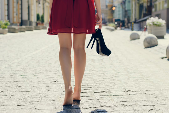 Woman in red dress, with high heel shoes in hand, walking in the city barefoot; close up photo of woman's legs, city on the background, view from back