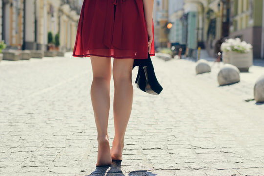 Elegant pretty woman with shoes in hands tired because of wearing high-heels walking along the street barefoot; close up photo of woman's long barefoot legs, view from back