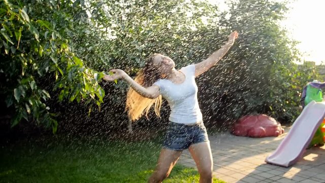 Slow motion video of happy cheerful young woman with long hair dancing under summer rain at backyard