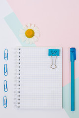 Top view photo of school supplies. Set of open empty notebook, pen, paper clips and chamomile on pink and blue background