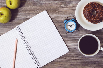 Stationery background. Opened notepad, alarm clock, plate with donut on it, cup of hot black coffee, apples on woode table