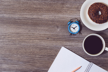 Set of modern workplace, alarm clock, opened notepad, cup of hot black coffee, donut.