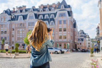 Happy, excited, stylish young woman taking photo of landmark in European city using her professional photo camera. She is dressed in jeand and shirt, she has lond blonde hair. View from back
