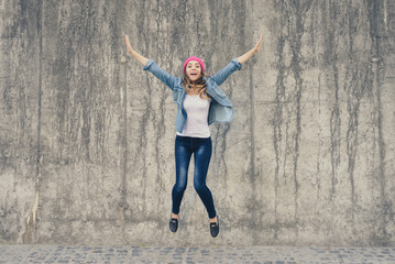 Obraz na płótnie Canvas Concept of joy and freedom, life without problems. Crazy, extremely happy girl in jeans clothes and pink hat screaming and jumping with raised hands against grey background.