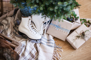 Gifts, presents and white children skates are under natural Christmas tree on wooden warm floor and beige cozy plaid. Blue decorating in rustic style with kraft paper, natural materials.