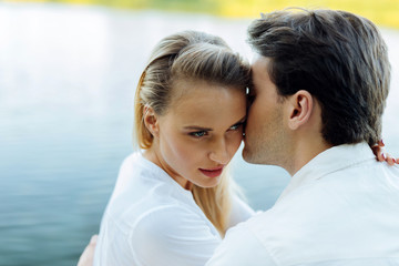 Fototapeta premium Happy couple. Beautiful young woman being kissed by her husband while sitting near the river
