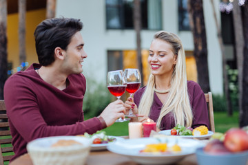 To us. Happy nice couple sitting together at the table while clinking glasses with wine