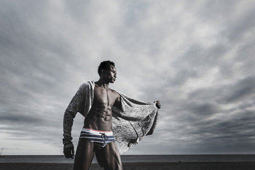 fashion and trendy concept for handsome black race afro model with swimsuit and jacket posing at the beach with ocean and blue sunset sky in background. Cold metal filter colors