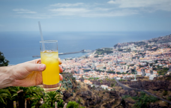 Drinking orange cocktail of Poncha on Madeira island, man holding a glass with drink, blurred city of Funchal on the background. Poncha is a traditional alcoholic drink from the island of Madeira.