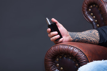 Hand close up. Vaper. The man with tattoos sits on a leather sofa smoke an electronic cigarette on...