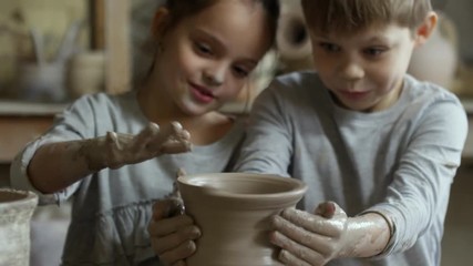 Tilt up of adorable elementary school-age girl and boy trying to make pottery on spinning wheel: they are throwing vase and laughing after failing and damaging it