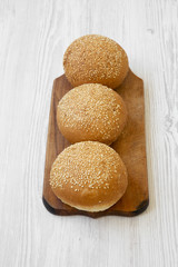Fresh burger buns with sesame seedson wooden board over white wooden background, high angle view. Fast food.