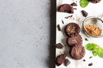 Chocolate cookies, pieces of chocolate, mint and cocoa powder on a white wooden cutting board. 