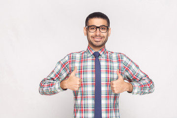 portrait of happy satisfied handsome bearded businessman in colorful checkered shirt, blue tie and black eyeglasses standing with thumbs up and toothy smile. studio shot, isolated on grey background.