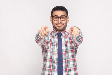 Hey you. portrait of happy bearded businessman in colorful checkered shirt, blue tie and black eyeglasses standing and pointing at camera with smile. studio shot, isolated on light grey background.