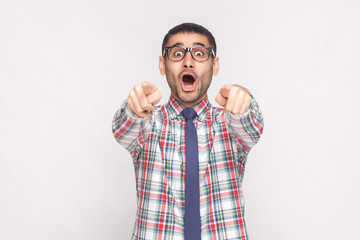 Amazed bearded businessman in colorful checkered shirt, blue tie and black eyeglasses standing, pointing and looking at camera with wondered open mouth. studio shot, isolated on light grey background