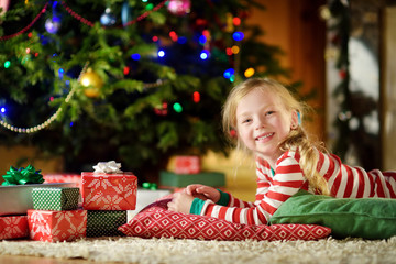 Happy little girl wearing Christmas pajamas playing by a fireplace in a cozy dark living room on Christmas eve.