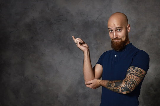 Look at that! Isolated studio shot of funny bald bearded man with tattoo raising index fingre and pointing at left corner, expressing excitement or curiosity, raising eyebrows. Body language