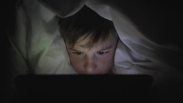 A happy boy lies in bed under a blanket and plays on a tablet in a game in the dark. The face of the child is lit by a bright monitor
