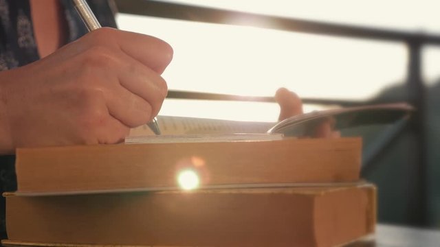 Woman's hand close-up, writing a note or letter to a notebook with a pen on a sunny day at sunset. A stack of scientific books, preparation for exams. Filling out the report
