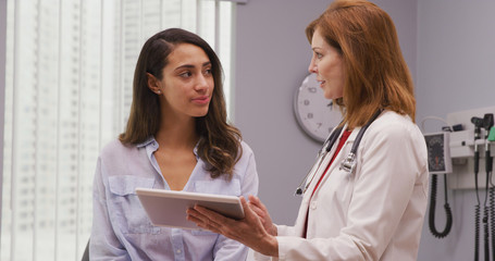 Portrait of lovely young hispanic woman meeting with senior doctor about pregnancy results. Close up of mid aged caucasian doctor using portable tablet to review health history with latina patient