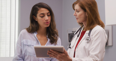 Camera pan of medical office with young hispanic patient and senior caucasian doctor looking at tablet device. Closeup of mid aged doctor using high tech tablet  to review health history with patient