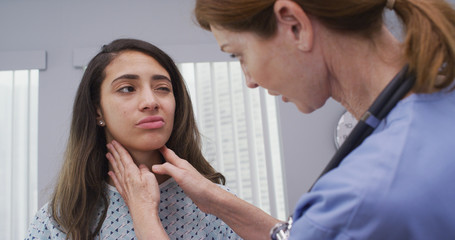 Medical nurse comforts young female patient holding her shoulder indoors hospital room. Mid aged nurse comforts young latina patient wearing hospital gown