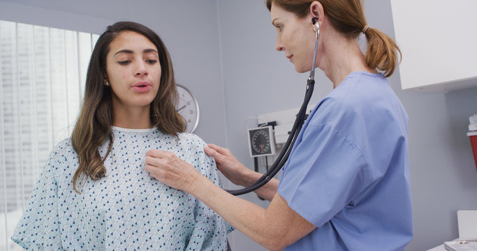 Mid Aged Nurse Using Stethoscope To Examine Young Patients Lungs And Heart. Young Latina Patient Takes Deep Breathes While Nurse Uses Medical Instrument To  Check Body Vitals