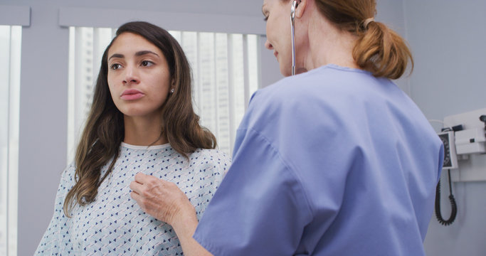 Close Up Of Lovely Latina Woman Breathing Deep Breaths While Nurse Examines Vital Organs. Young Patient Seated In Medical Clinic Taking Long Breaths As Mid Aged Nurse Uses Stethoscope