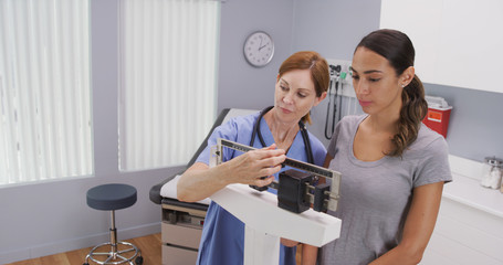 Mid aged caucasian nurse weighing female patient on scale. Charming middle aged nurse using scale to measure weight of young latina patient - obrazy, fototapety, plakaty