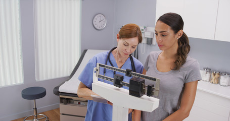 Mid aged caucasian nurse weighing female patient on scale. Charming middle aged nurse using scale to measure weight of young latina patient