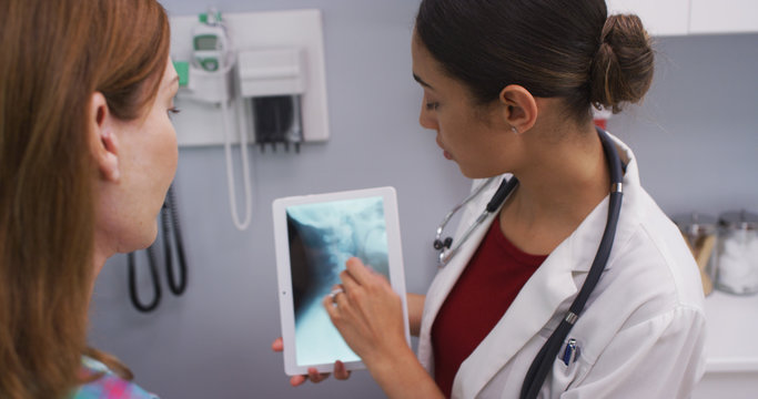 Portrait of young doctor using tablet to review x ray of female patients neck.  Close up of attractive latina doctor and senior patient looking at x-ray of spine and cranium