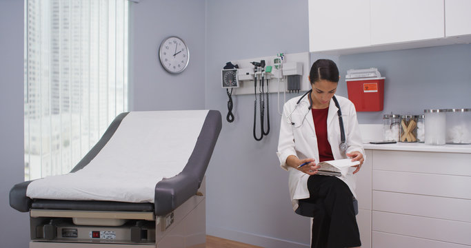 Close Up Of Lovely Latina Doctor Reviewing Patients File Indoors Medical Clinic. Attractive Young Doctor Checking Notes On Clipboard For Patients Health History