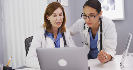 Intelligent young latina doctor looking at laptop computer and talking with elder physician. Two medical doctors using notebook computer to discuss patients healh condition
