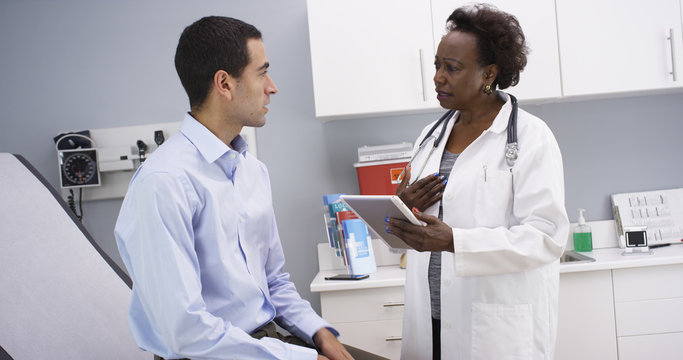 African senior medical doctor using high-tech elctronic notebook to take notes of patient. Portrait of young hispanic patient consulting with middle aged black doctor indoors medical clinic