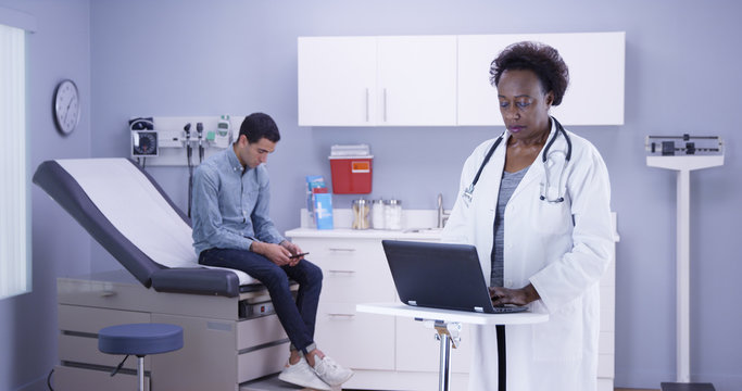 Wide Shot Of Medical Clinic With Mid Aged African Doctor Typing On Laptop While Young Male Patient Uses Smartphone. Senior Black Doctor Taking Notes Of Single Latino Patient On Portable Computer