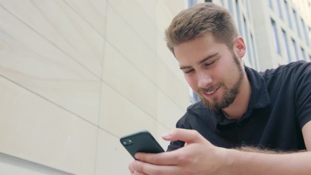 A man with a beard sitting in front of a modern building and using a phone. Close-up shot. Soft focus