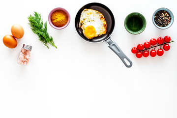 Cooking, preparing fried eggs with vegetables. Ready eggs in a frying pan near cherry tomatoes, greenery, spices, raw eggs on white background top view copy space