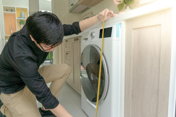 Young Asian worker man using tape measure for measuring washing machine in kitchen counter. Home...