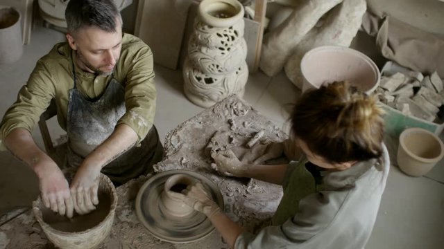 High angle shot with PAN of bearded man and young woman throwing pot on spinning pottery wheel in workshop