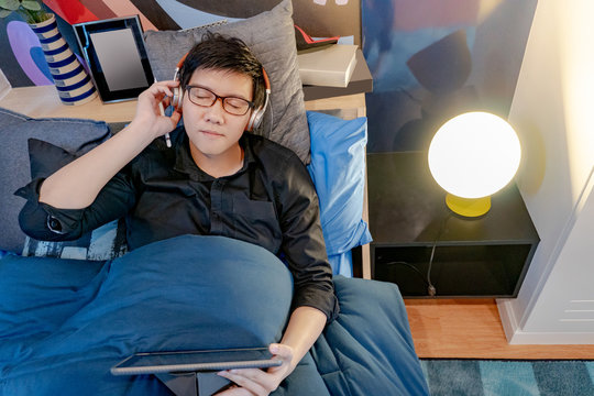 Young Asian happy man listening to music with headphones while using digital tablet and pen on his comfortable blue bed in the bedroom. Home living lifestyle with modern electronic gadgets concept